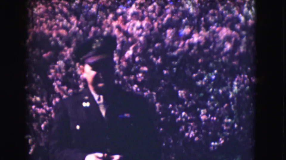 1975 Washington DC home-movie frame showing a uniformed man in a dark cap before a purple-tinged hedge.