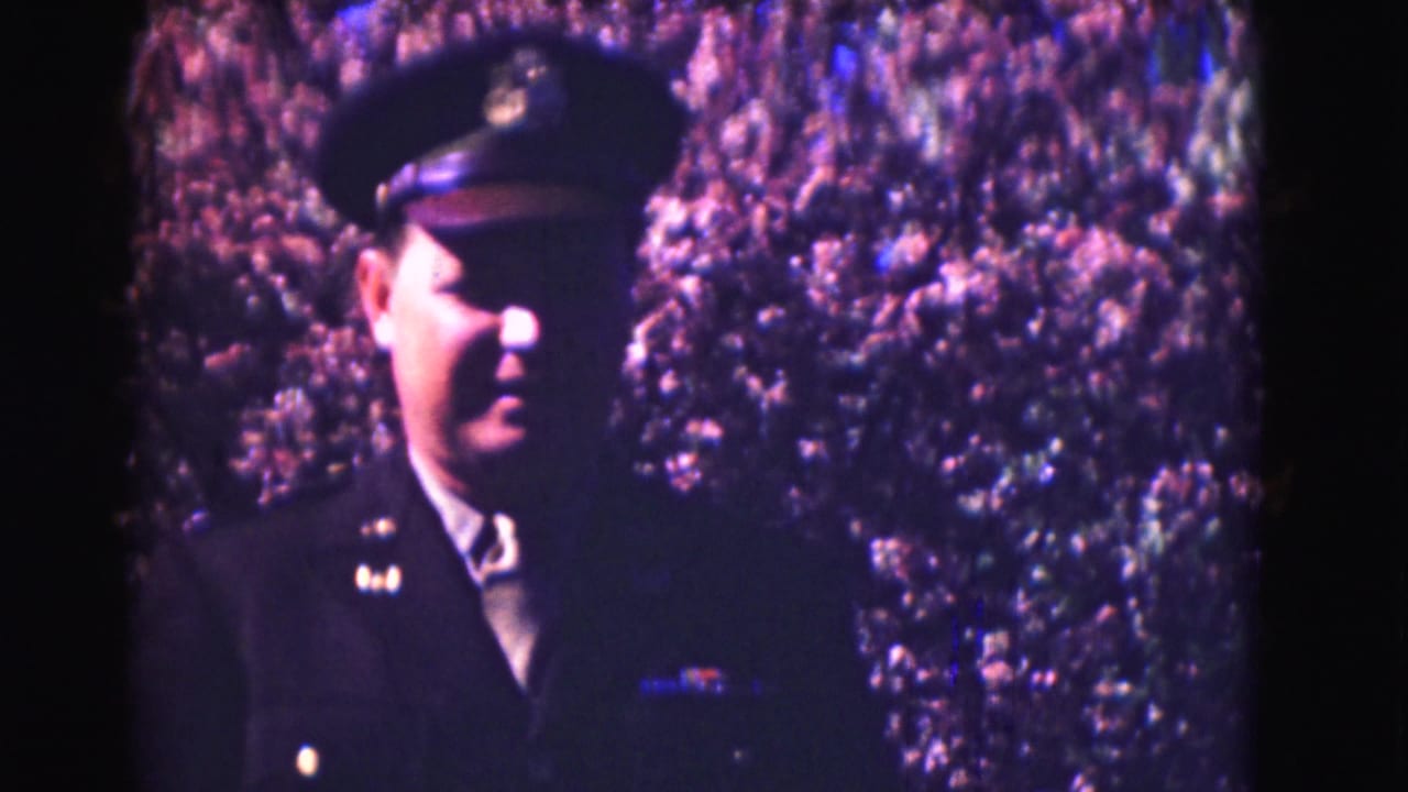 Closer view of a uniformed man in cap before flowering shrubs.