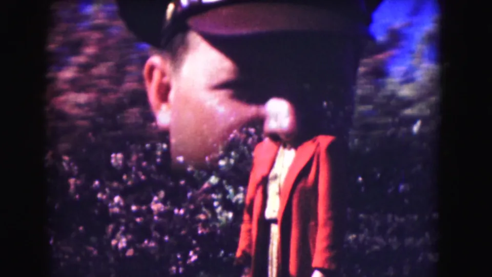 1975 Washington DC home-movie frame with a woman in a red coat and light top standing before dense shrubs.
