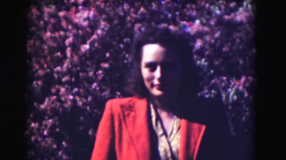 Home-movie close-up of a woman in a red jacket, with pink flowering trees filling the background.