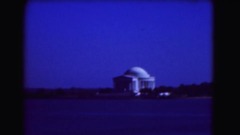 Distant 1975 Washington, DC view of the Jefferson Memorial glowing across the Tidal Basin, with a dark treeline below a blue sky.