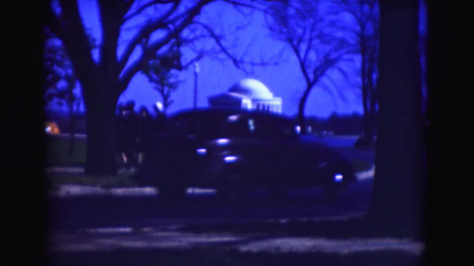 A blue-tinted 1975 Washington, DC frame shows a dark car crossing the foreground while visitors stand near the Jefferson Memorial grounds.