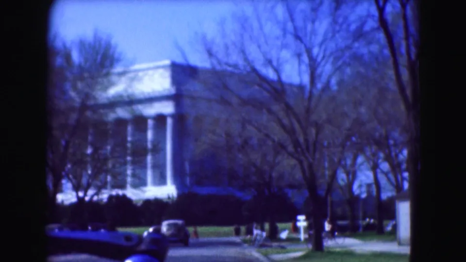 1975 Washington, DC 8mm frame with a columned building, bare trees, a blue car, and small distant pedestrians near the roadway.