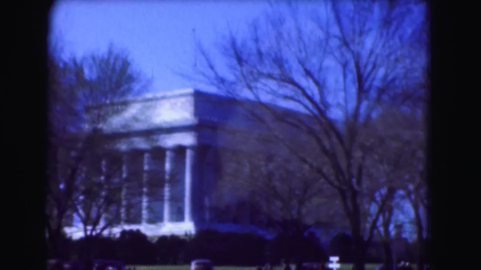 Soft 1975 home-movie frame from Washington, DC showing a columned facade behind leafless trees, with cars along the lower foreground.