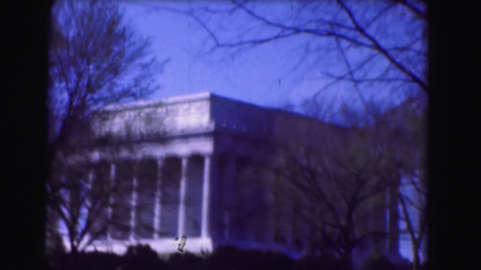 Blue-shifted 1975 home-movie footage shows the Lincoln Memorial partially screened by trees and foreground shrubs.
