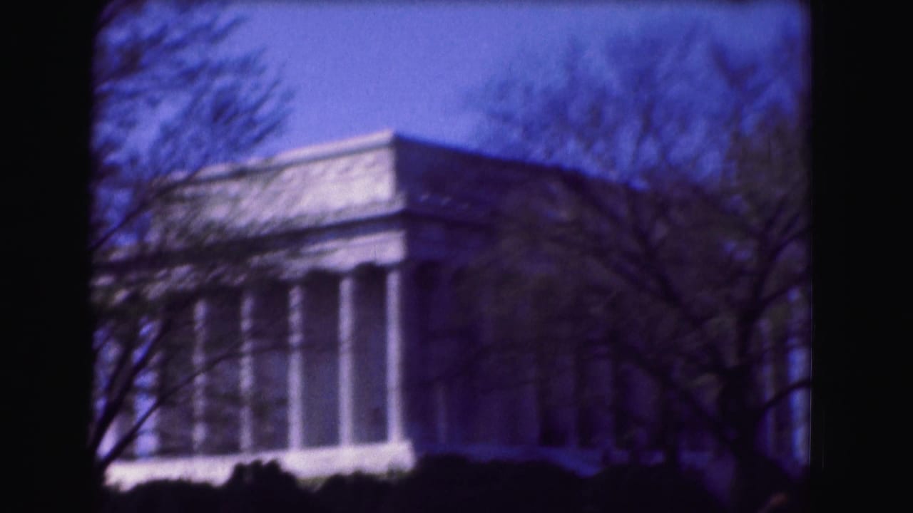 1975 Washington, DC home-movie frame showing the Lincoln Memorial facade partly obscured by bare branches and dark foreground shrubs.