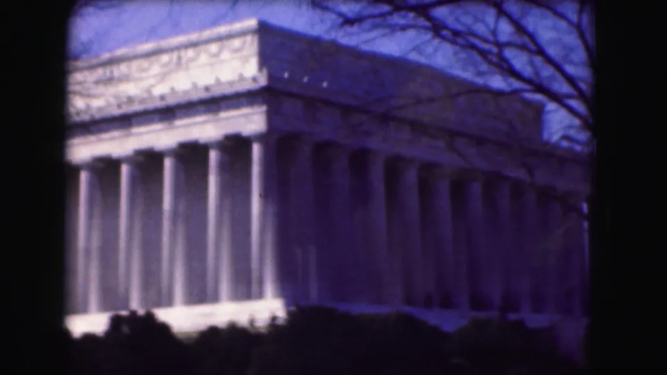 1975 Washington, DC frame showing the Lincoln Memorial from an angle, with its columned side, dark shrubs, bare trees, and blue sky.