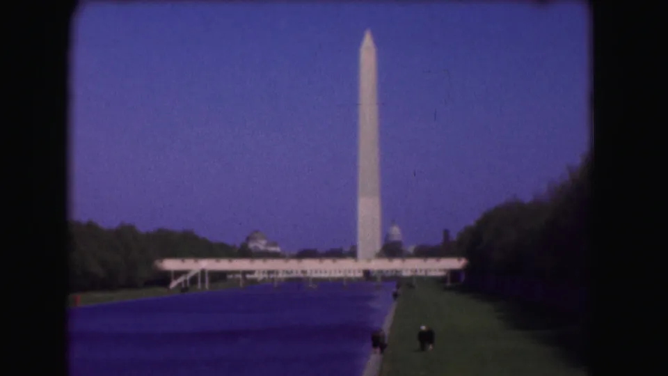Washington Monument rises beyond the Reflecting Pool on the National Mall, with lawns, trees, distant visitors, and the Capitol dome faintly visible.