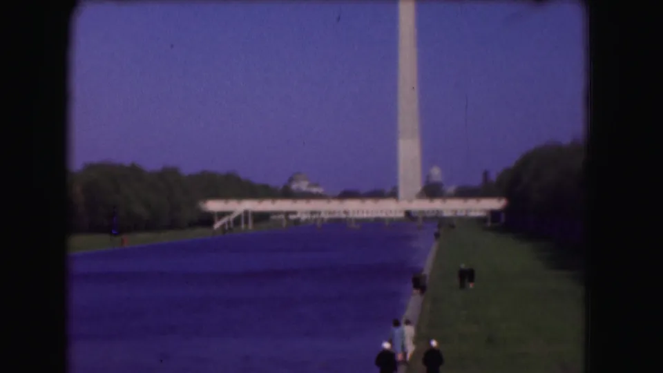Handheld 8mm frame of the Reflecting Pool leading toward the Washington Monument, with lawns, trees, and small visitors along the right edge.