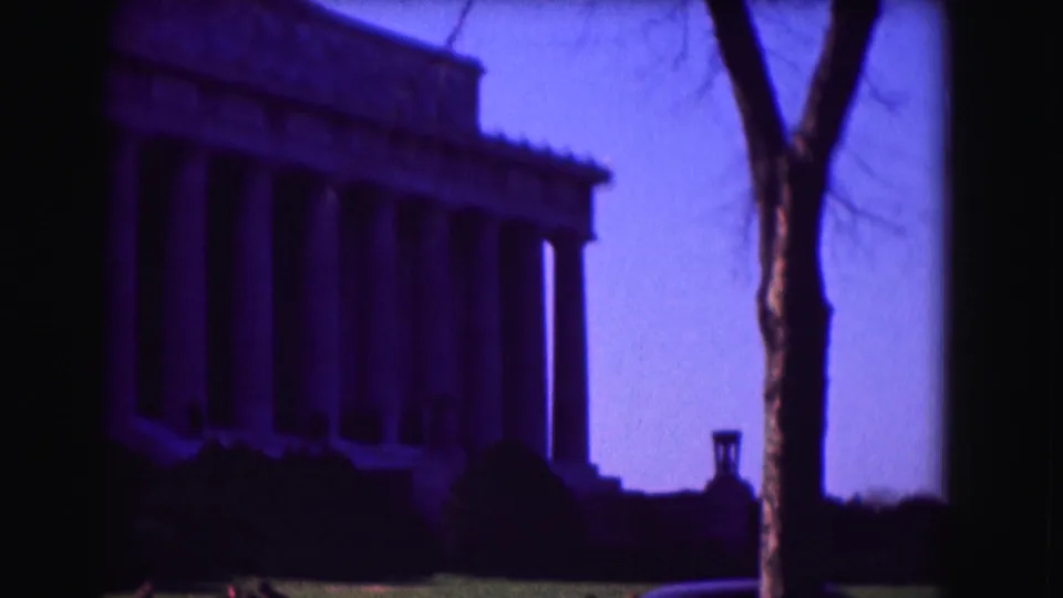 1975 8mm Washington, DC frame showing the Lincoln Memorial behind a bare tree trunk, with lawn, shrubs, and a distant visitor near the base.