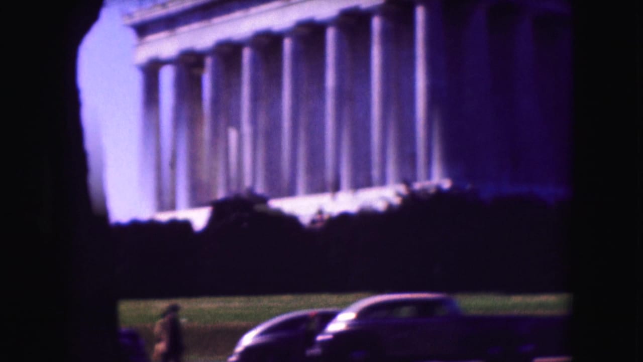 Washington, D.C. home-movie frame showing the Lincoln Memorial facade, lawn, a dark car, and distant visitors.