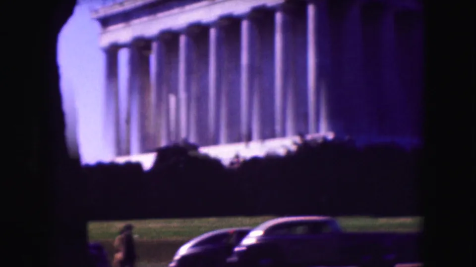 Washington, D.C. home-movie frame showing the Lincoln Memorial facade, lawn, a dark car, and distant visitors.