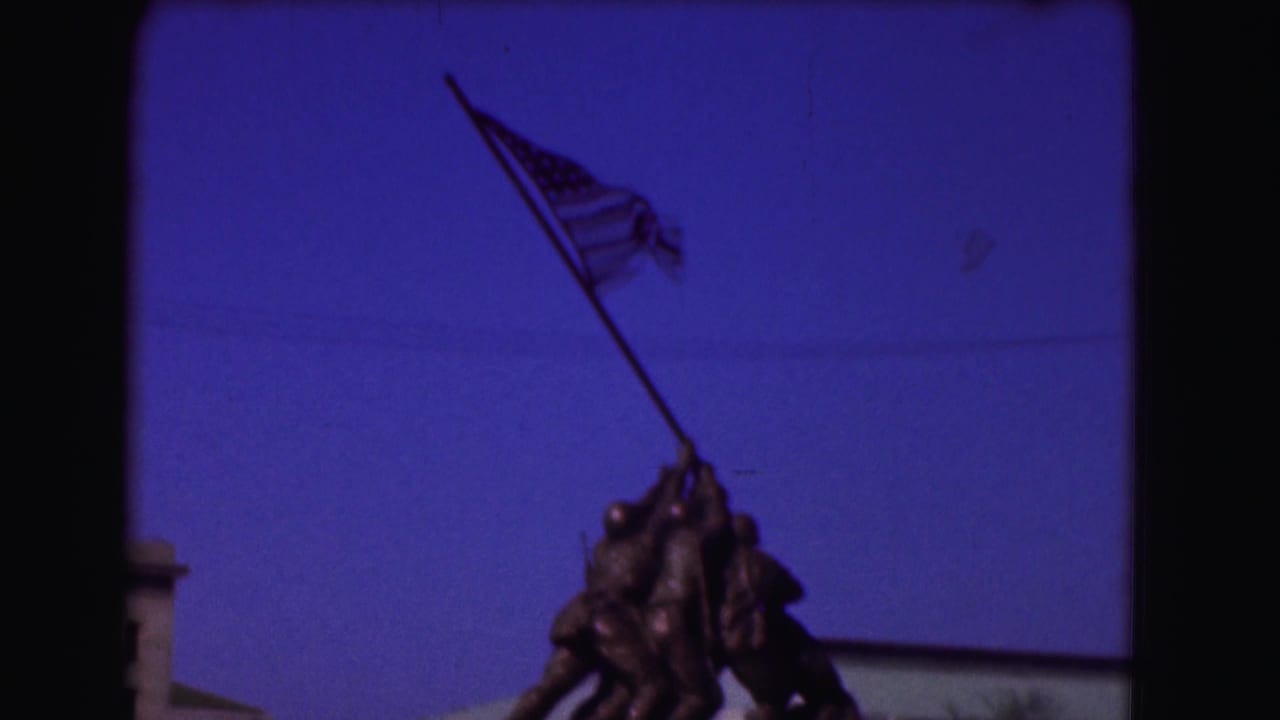 1975 home-movie frame of the Iwo Jima Memorial, with the American flag and upper bronze figures under a clear blue sky.