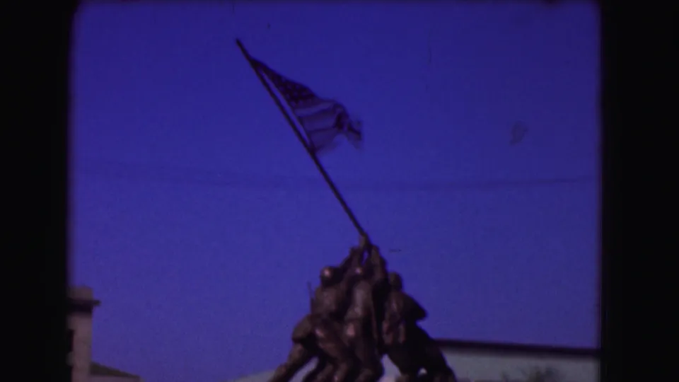1975 home-movie frame of the Iwo Jima Memorial, with the American flag and upper bronze figures under a clear blue sky.