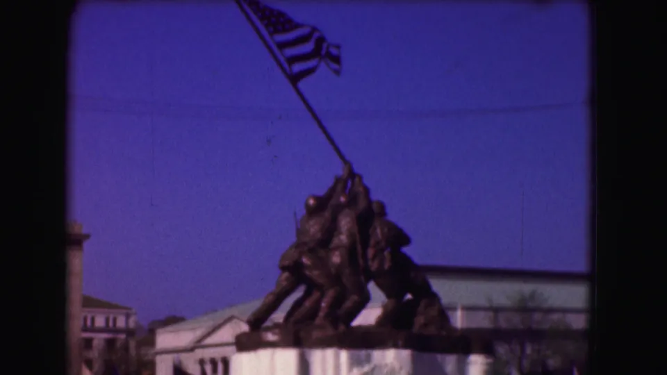 1975 8mm close-up of the Iwo Jima Memorial figures lifting the flag, with nearby architecture at the frame edges.
