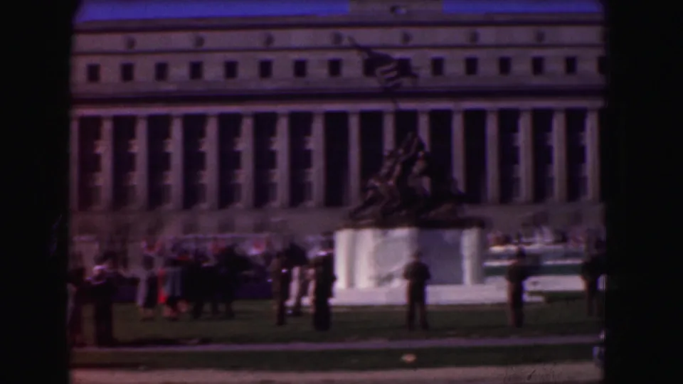 1975 Washington, DC wide 8mm frame showing a war memorial statue on a plaza with visitors and a large classical building in the background.