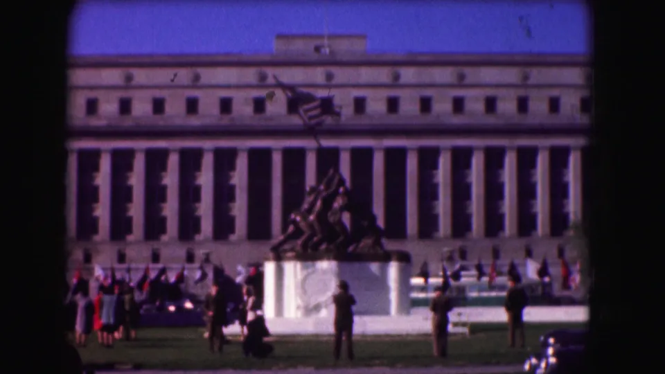 1975 Washington, D.C. 8mm frame with the Iwo Jima statue, clustered visitors, flags, and personnel before a formal building.