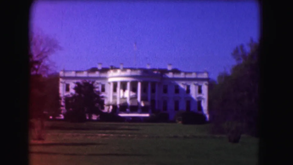 1975 8mm frame of the White House across the lawn, with trees, blue sky, and a reddish film-edge flare.
