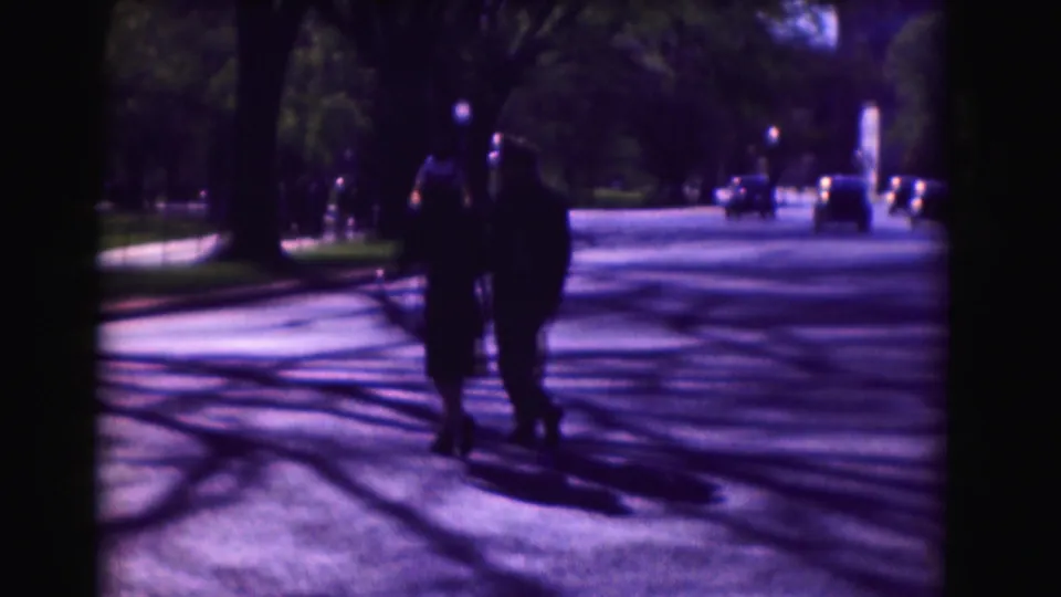 Two adults walk on a tree-lined Washington, DC street, with long shadows, parked or moving cars, and soft 8mm color.