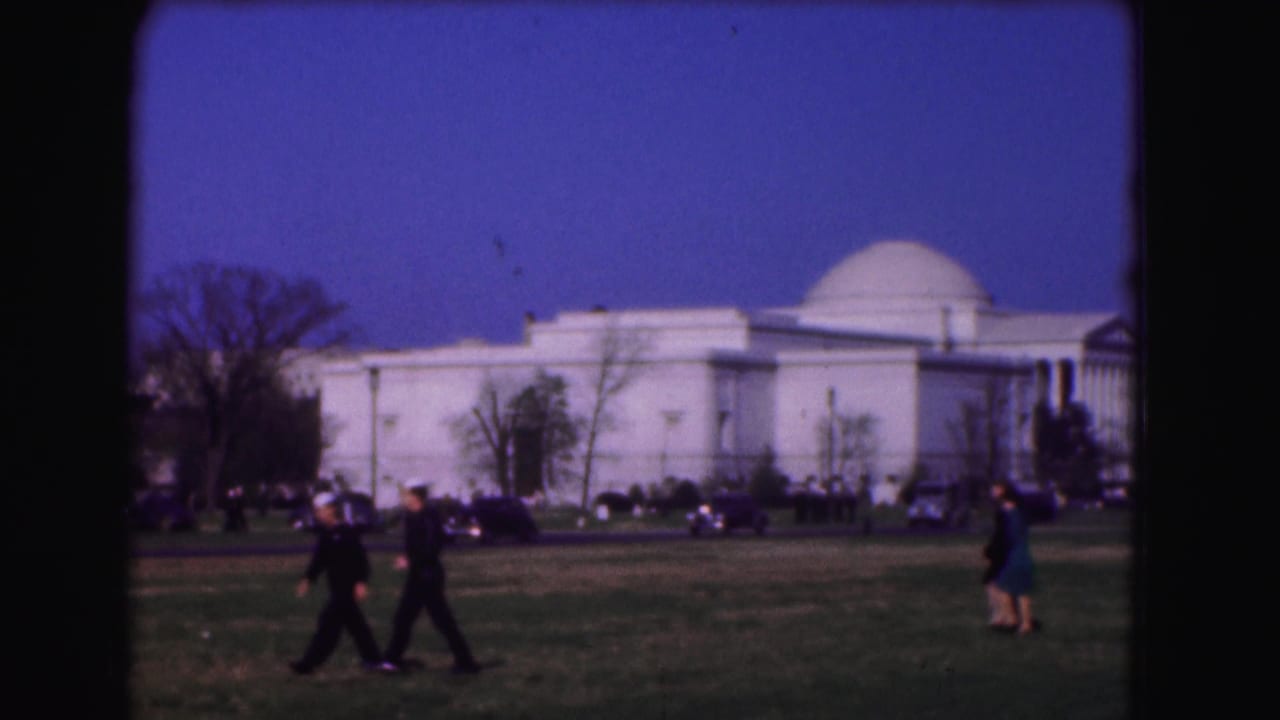 1975 Washington, DC frame with two pedestrians crossing a lawn in front of a white domed public building and parked cars.
