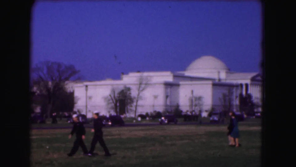 1975 Washington, DC frame with two pedestrians crossing a lawn in front of a white domed public building and parked cars.
