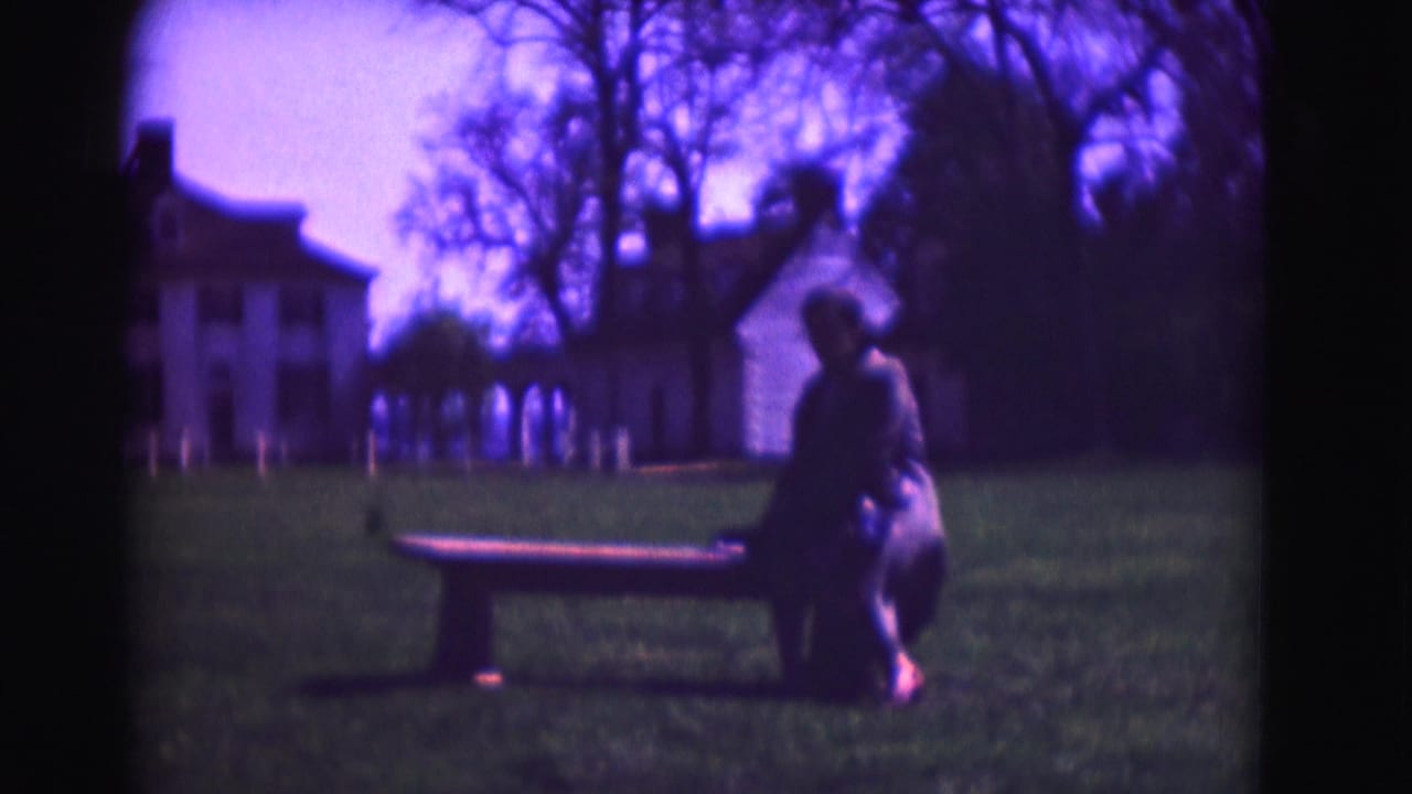 1975 Washington DC frame of a woman seated on a bench in a grassy estate setting, with a large white house in the background.