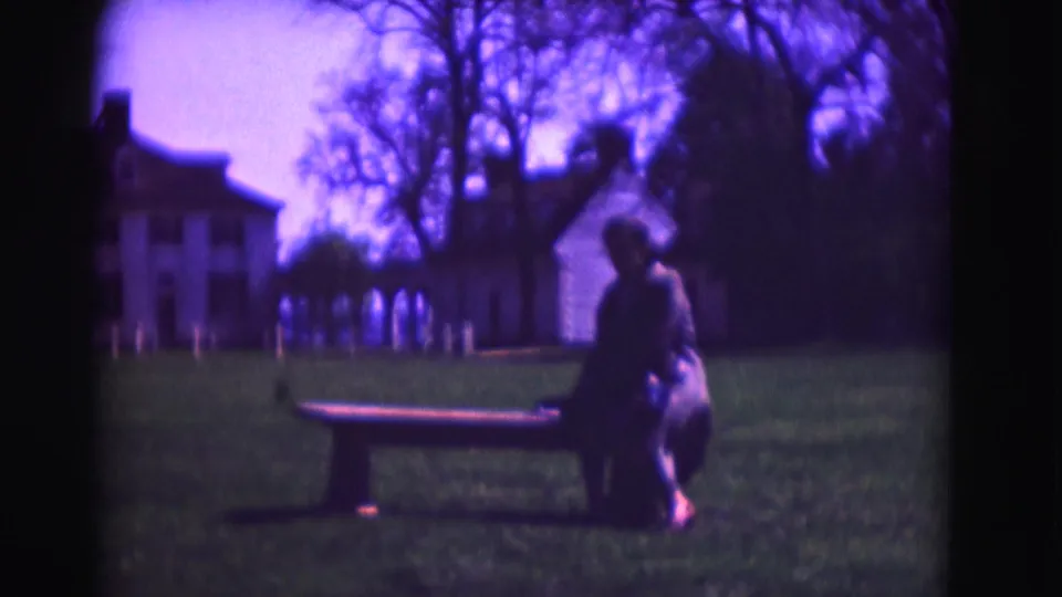 1975 Washington DC frame of a woman seated on a bench in a grassy estate setting, with a large white house in the background.