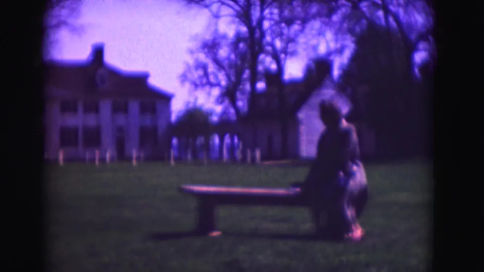 1975 Washington DC home-movie still showing a woman in a dark coat seated by a bench, with a white mansion and trees across the lawn.