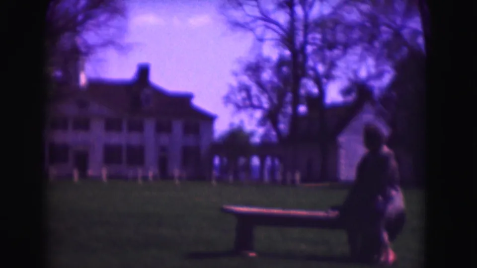 1975 Washington, DC home-movie frame showing a person on a wooden bench before white colonial-style houses and bare trees.