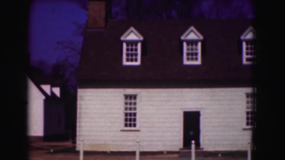 1975 Washington, DC home-movie frame showing a white clapboard house with dormers, a dark roof, bare trees, and white posts in front.