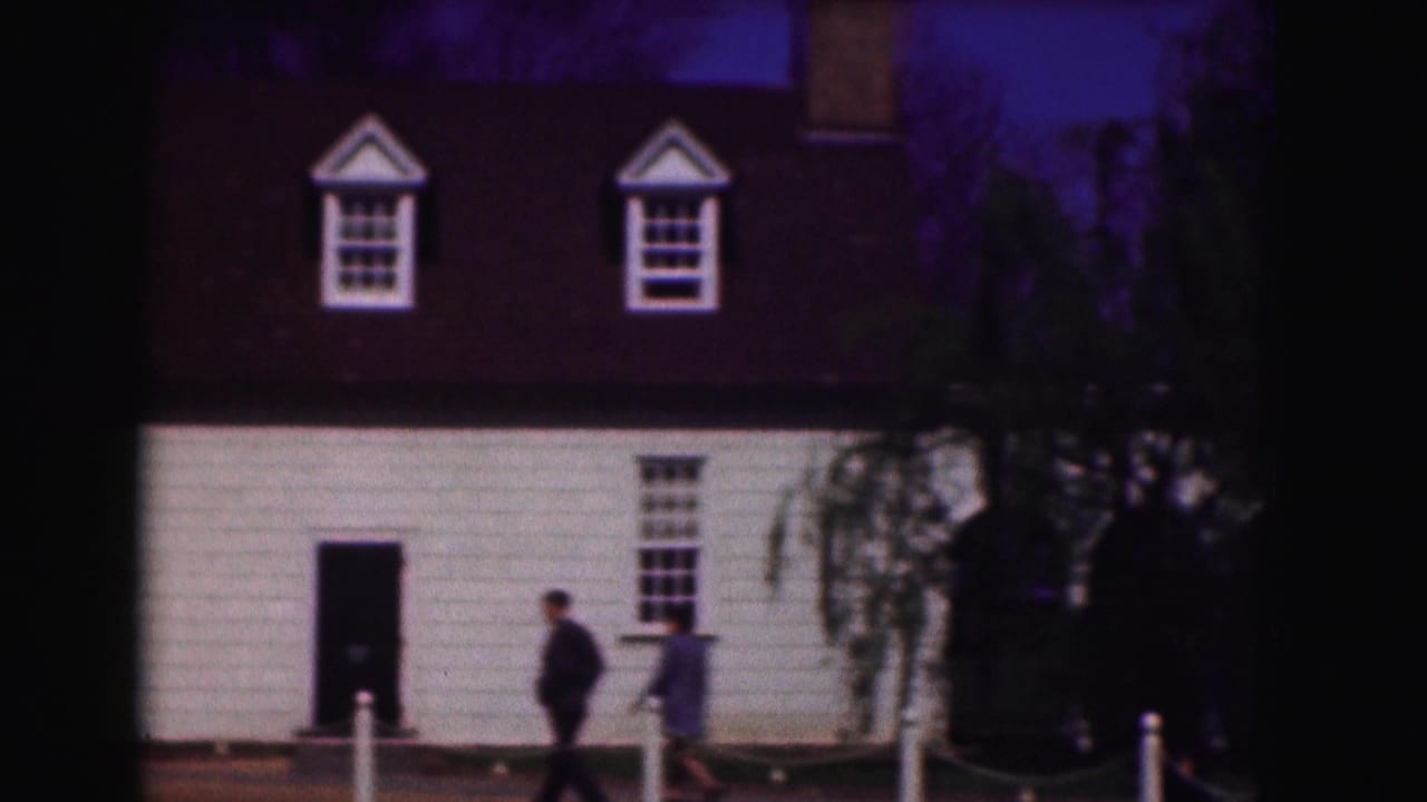 1975 Washington, DC frame with a person walking past the white clapboard building, dormer windows, doorway, trees, and foreground posts.