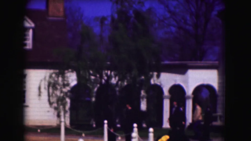 1975 Washington, DC home-movie frame showing an arched colonnade, trees, visitors in shadow, white posts, and part of the clapboard building.