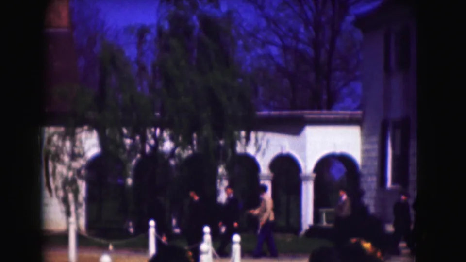 1975 Washington, DC frame with several visitors moving near arched openings, trees, white posts, and the edge of a clapboard structure.