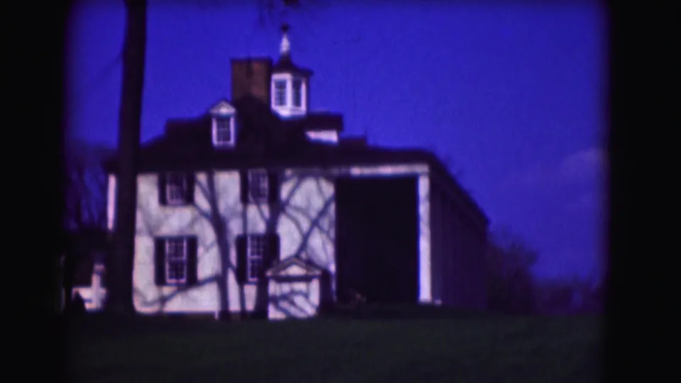 1975 8mm Washington, DC frame showing a white residence with cupola, dark shutters, lawn, and a person partly visible at left.