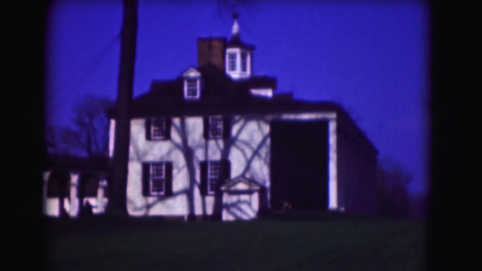 1975 8mm frame from Washington, DC showing a white two-story house with cupola, dark shutters, porch, lawn, and trees.