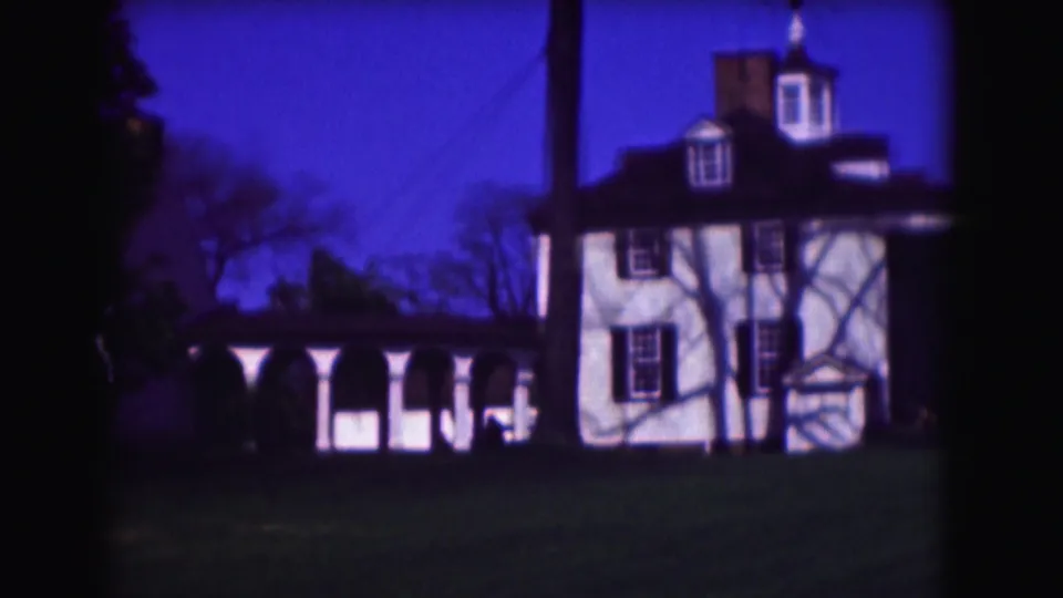 1975 Washington, DC home-movie frame showing the house facade, porch columns, lawn, trees, and strong blue film color.