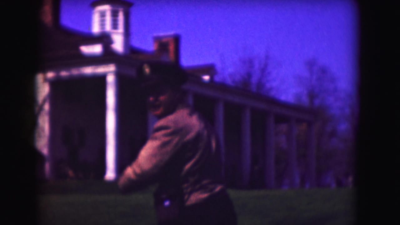 Uniformed man seen from behind on a lawn before a white columned building.