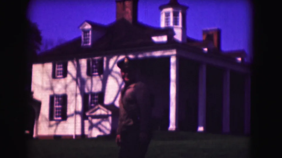 1975 8mm frame of a uniformed man in profile on a lawn before a white colonial-style building in Washington DC.