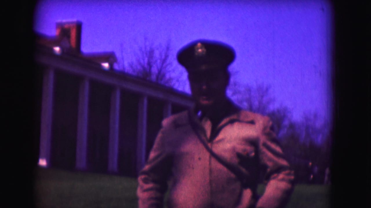 Uniformed man in a peaked cap standing on a lawn before a columned building.