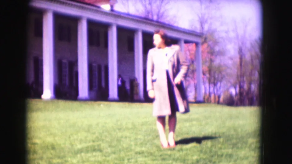 1975 Washington, DC home-movie frame of a woman in a long coat walking on grass near a large columned exterior.