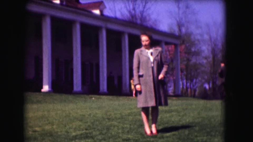 1975 Washington, DC frame of a woman in a long coat facing the camera on a lawn, with columns and trees in the background.