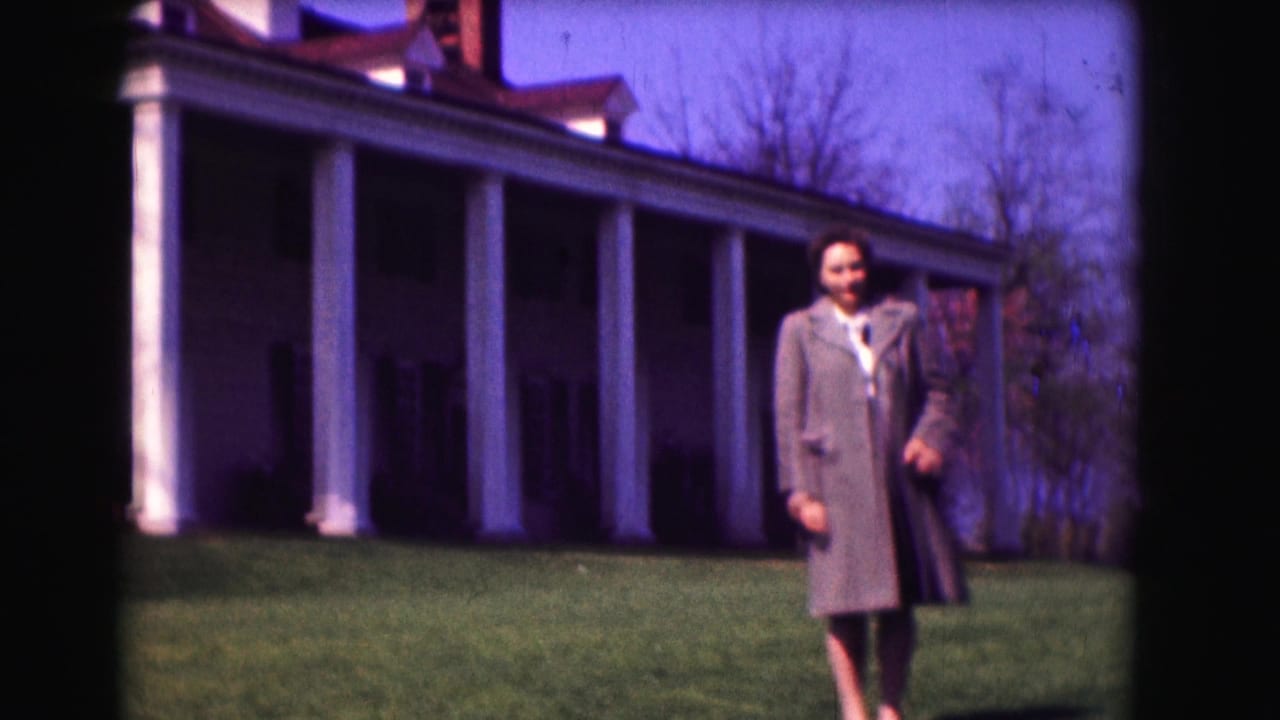 Woman in a gray overcoat standing on a lawn before a white columned house.