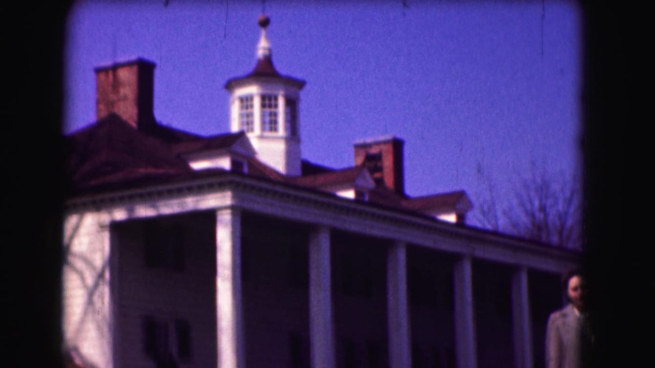 The camera frames the white historic-looking house, cupola, roof, and chimneys while the woman remains partly visible at right.