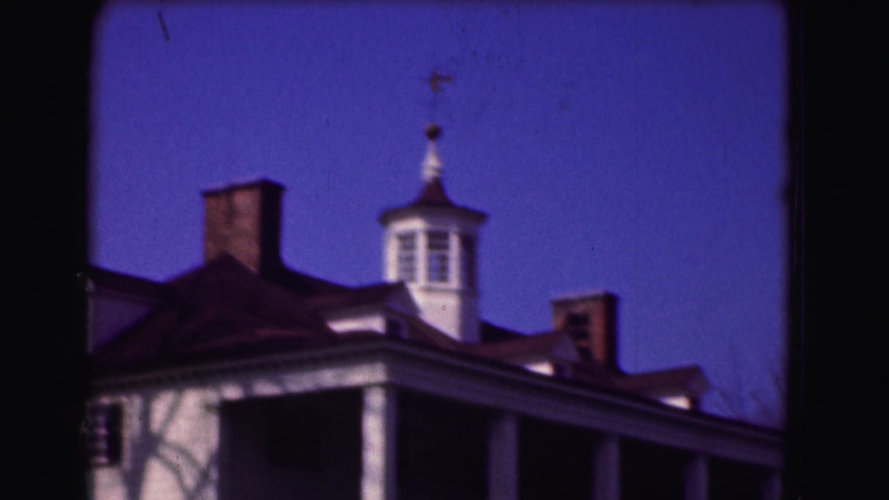 Colonial-style building roofline with white columns, brick chimneys, cupola, and blue sky.