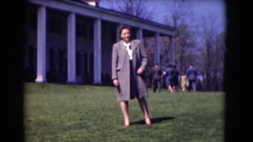 The woman is centered on the lawn in a 1975 Washington DC home-movie frame, with a columned facade and strolling visitors behind her.