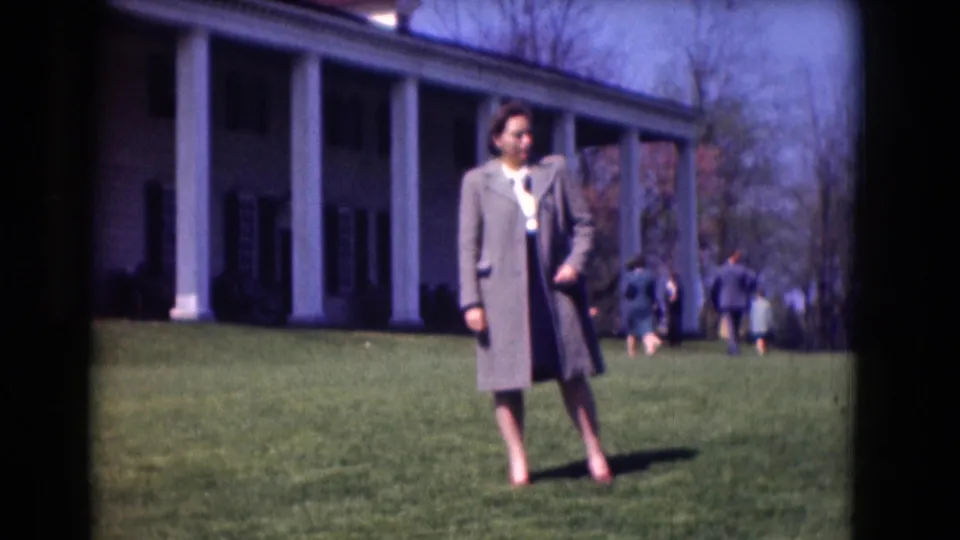 A woman stands on a sunlit lawn before a white-columned historic building, with a small visitor group crossing behind her.