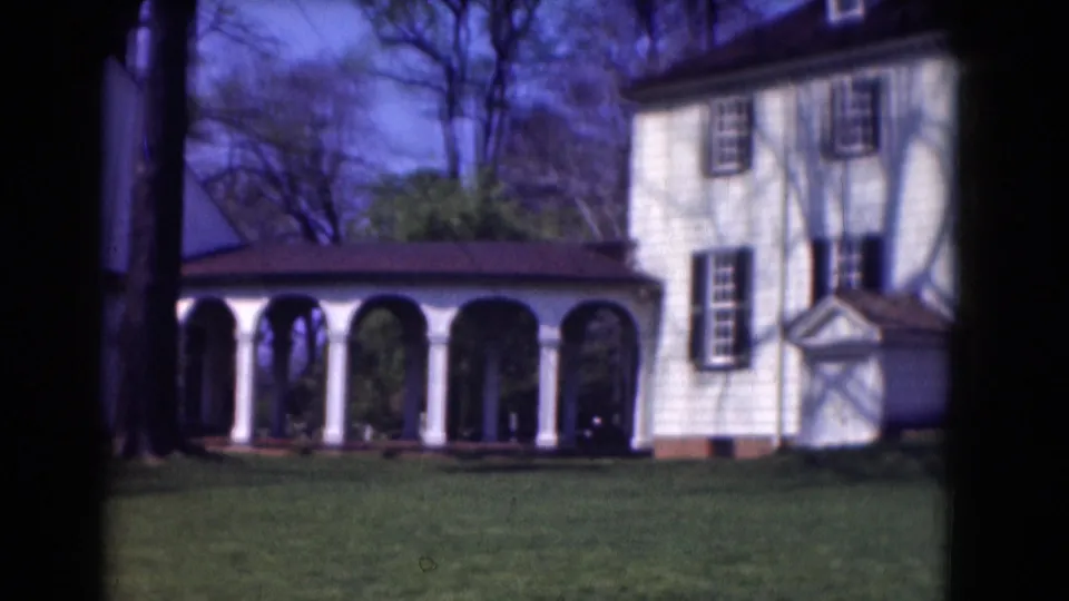 A white building with shuttered windows and an arched colonnade appears across a manicured lawn in 1975 home-movie footage.