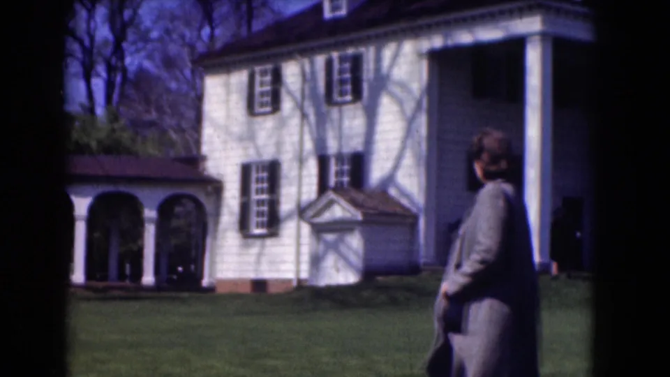 A woman in a gray coat walks at the edge of the frame beside a white house and arched walkway in a Washington DC 1975 home movie.