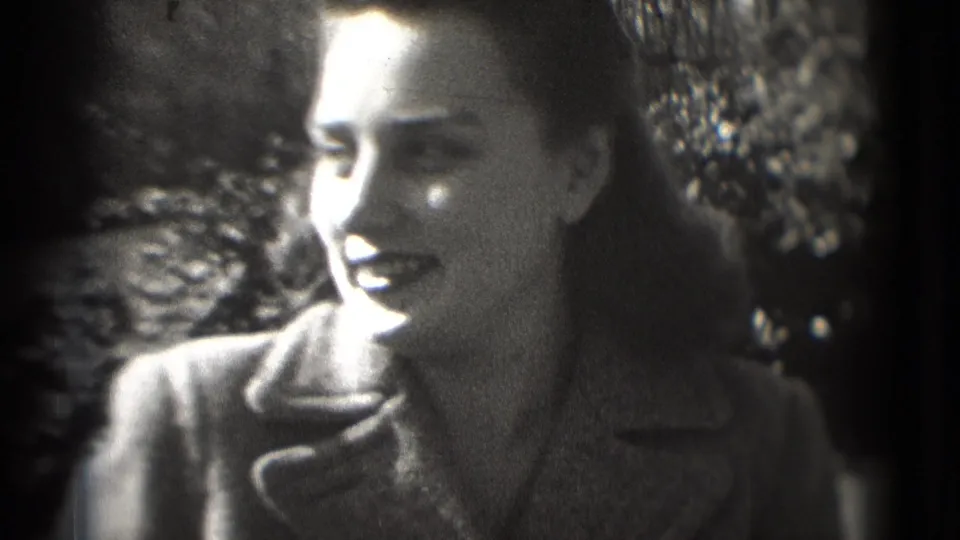 1975 8mm home-movie frame shows a woman in a dark coat and lipstick smiling outdoors, with trees behind her.