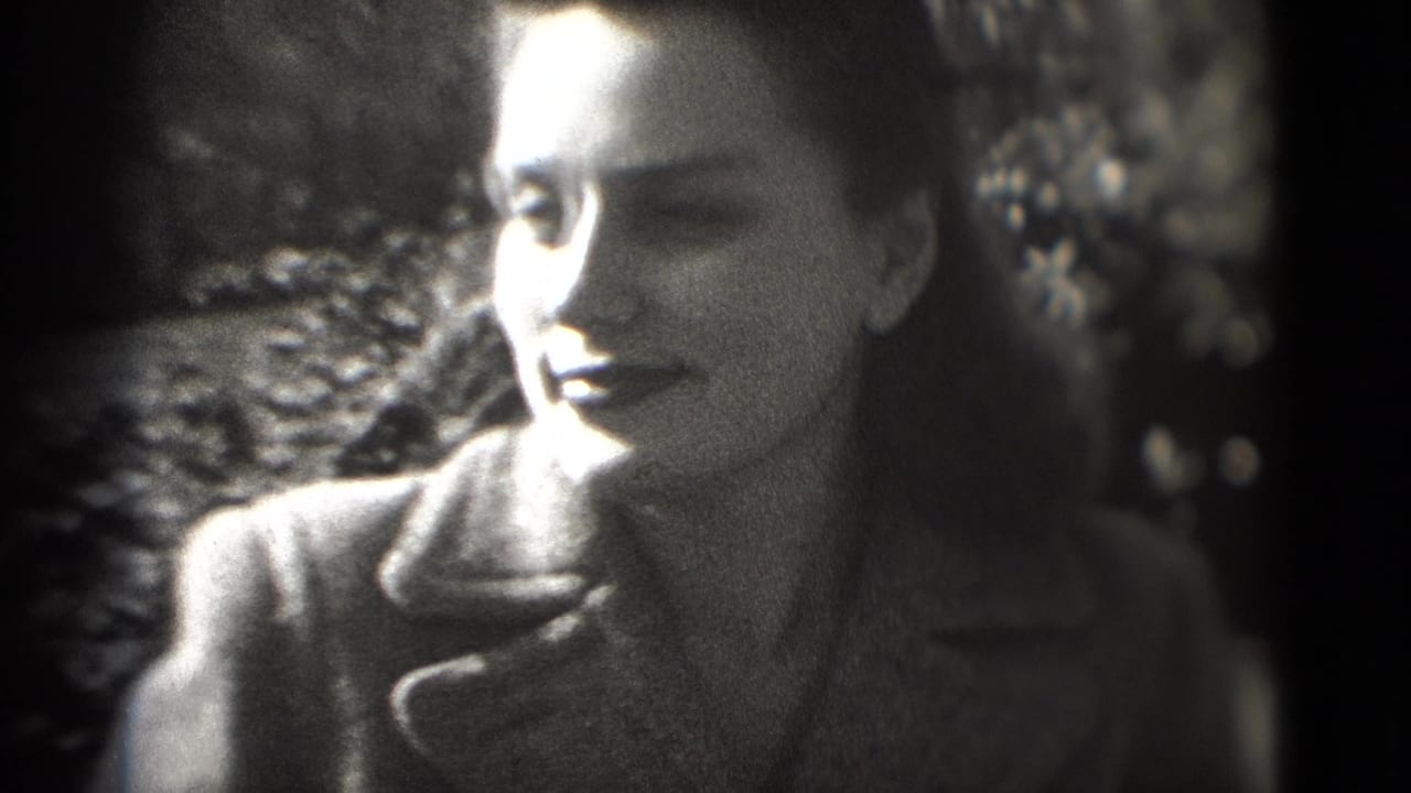 1975 8mm frame shows the woman turning slightly in profile, smiling in a dark coat against a leafy outdoor background.